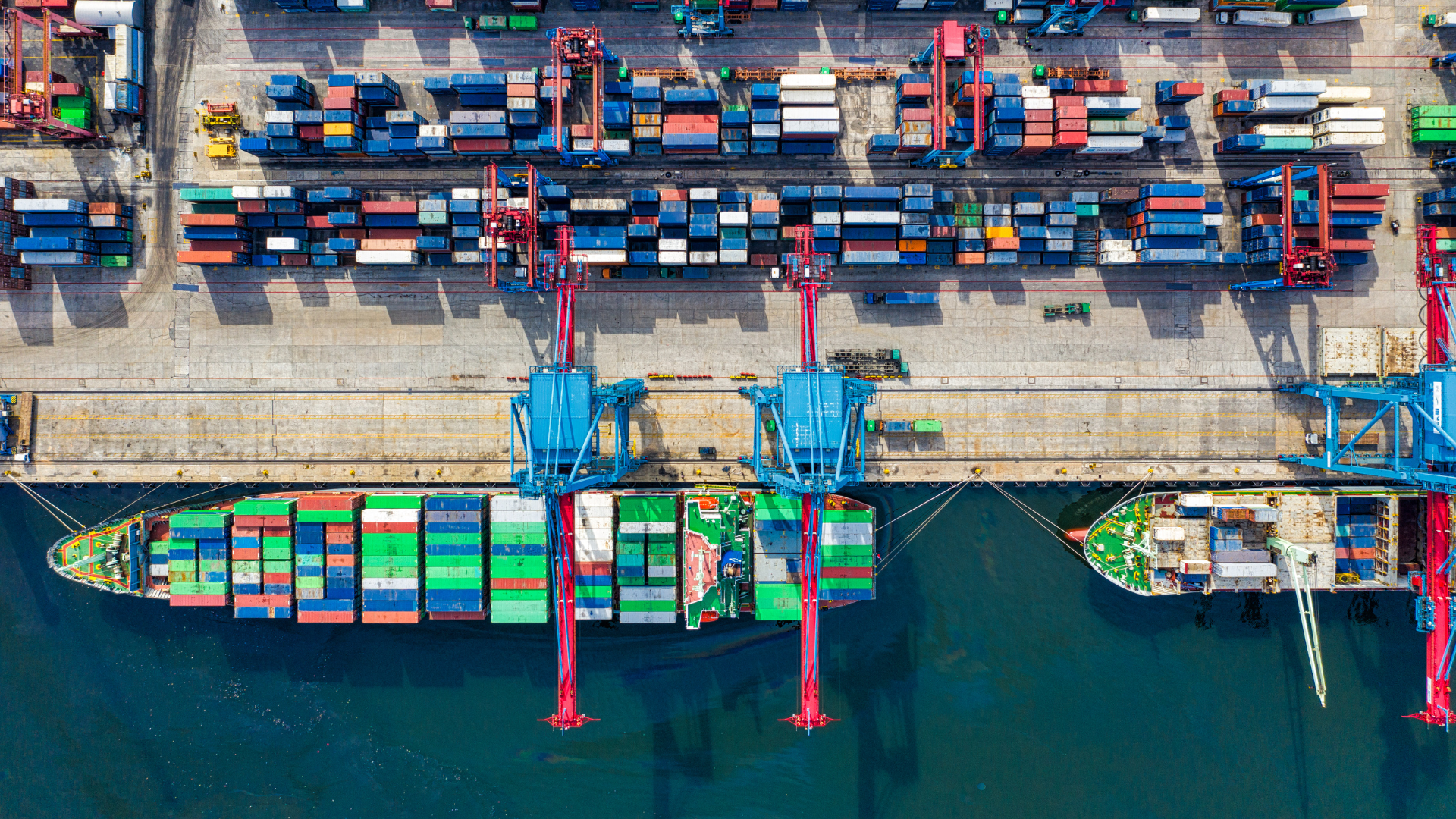 Vista aérea de um porto com navios cargueiros atracados, guindastes e contêineres organizados no terminal logístico, representando as mudanças no cenário internacional e seus impactos nas cadeias globais de suprimentos, comércio exterior e logística.